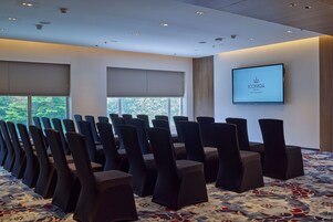 A conference room with rows of chairs, a large screen displaying ICONIQA HOTEL, and windows with blinds.