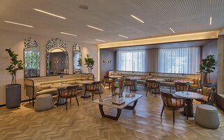 A spacious lounge area with a herringbone wood floor, a central coffee table, multiple seating options including sofas and chairs, and large windows providing natural light.