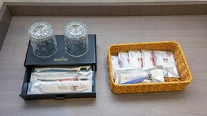 A hotel room with a black tray table, two crystal glasses, a black box with toiletries, and a wicker basket with towels.