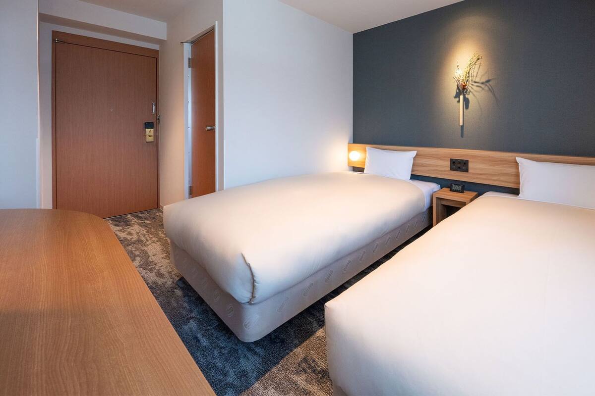 Ein Hotelzimmer mit zwei Betten, einem Holzkopfstück, einer wandmontierten Lampe und einer Tür.