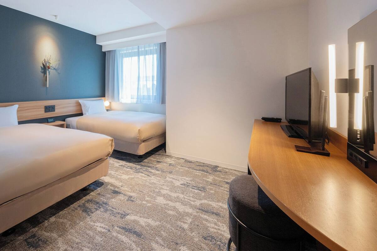 Ein Hotelzimmer mit zwei Betten, einem Schreibtisch, einem Flachbildfernseher, einer Wandlampe und einem Fenster mit Vorhängen.