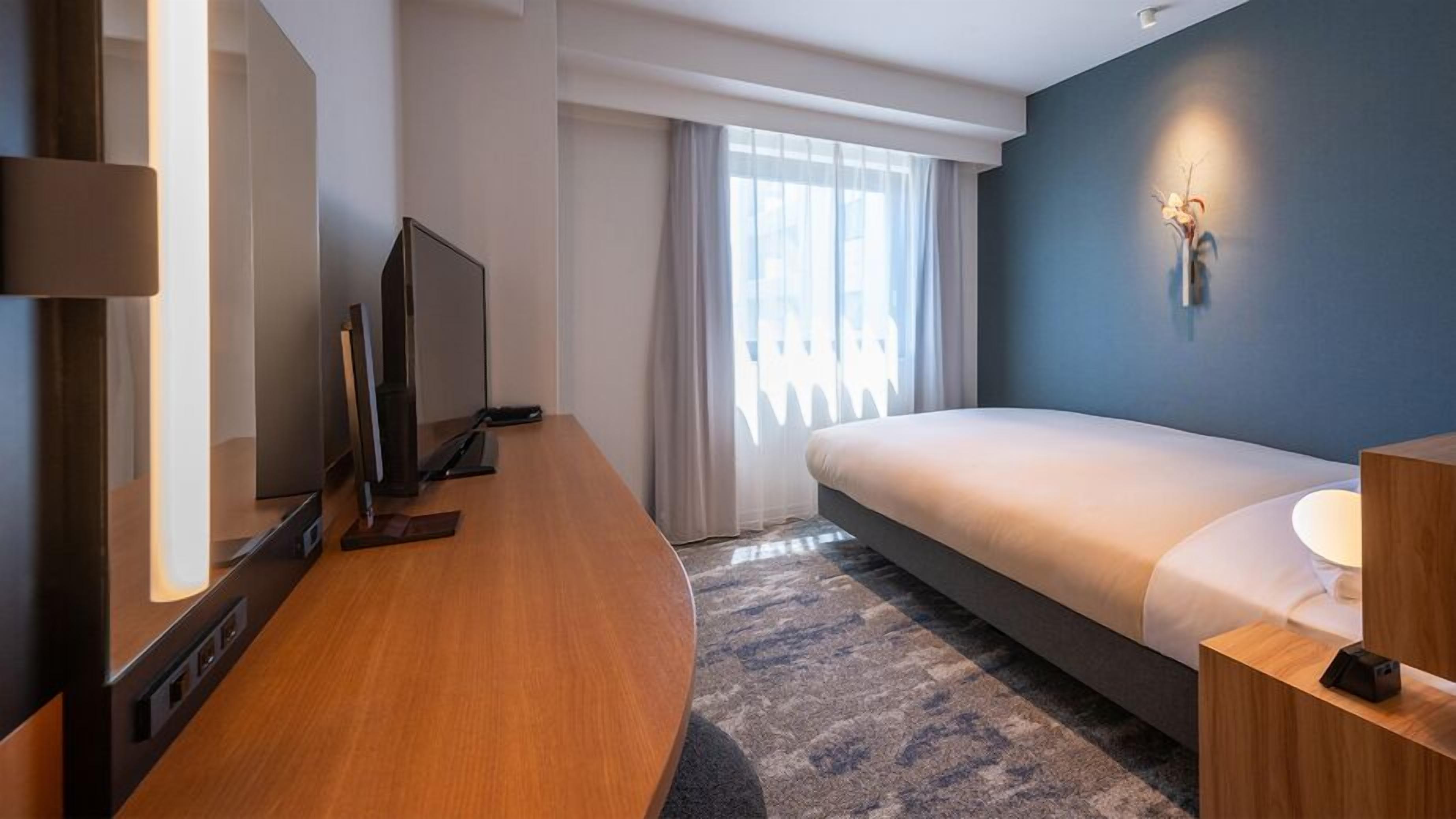 Una camera d'albergo moderna con un letto, una scrivania, una televisione e una lampada a parete.