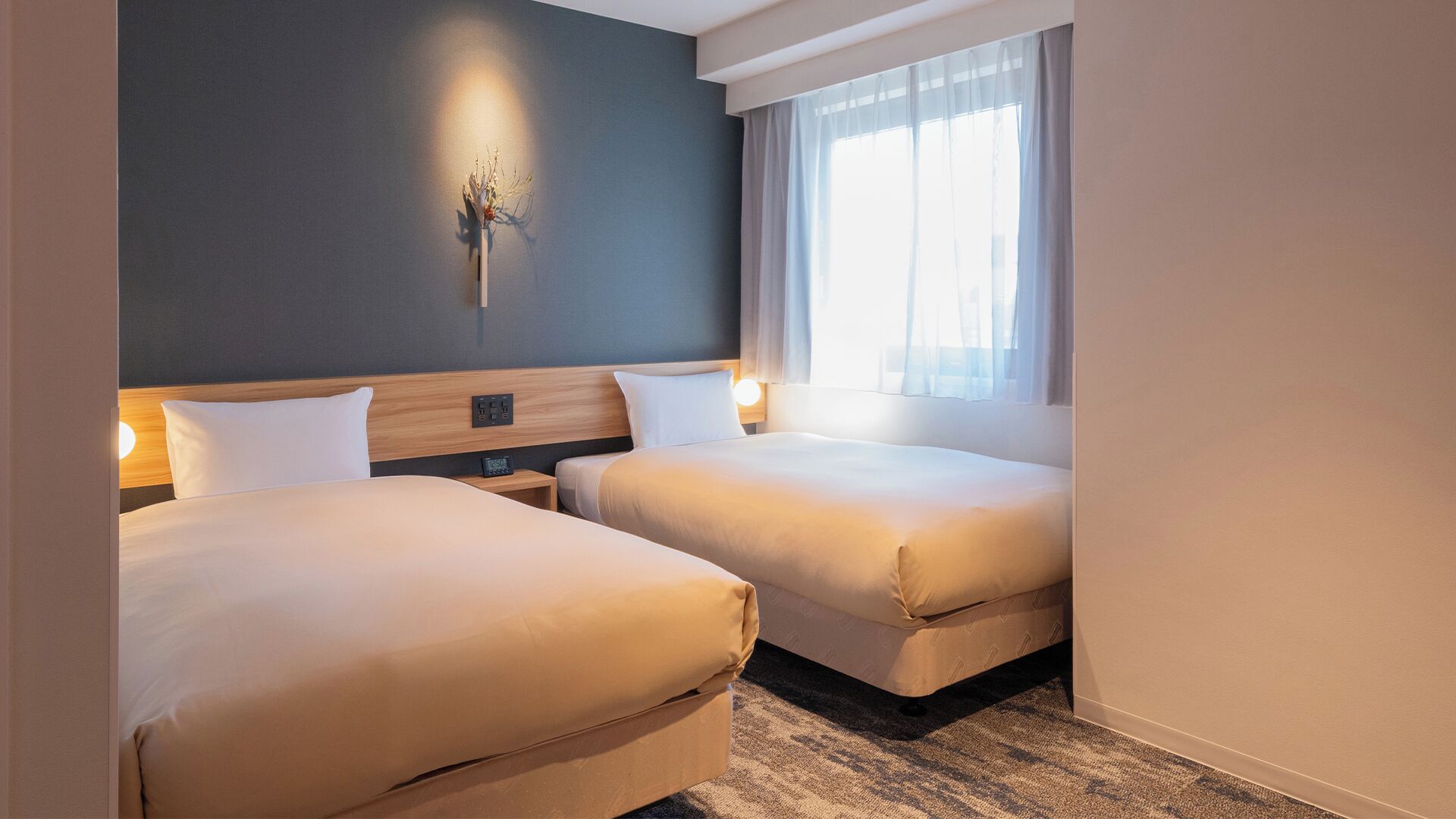 Habitación de hotel con dos camas, una pared oscura, una ventana con cortinas traslúcidas y una lámpara fijada a la pared.