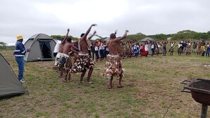 Een groep mensen in traditionele kledij danst in een open veld, met tenten en een barbecue op de achtergrond.