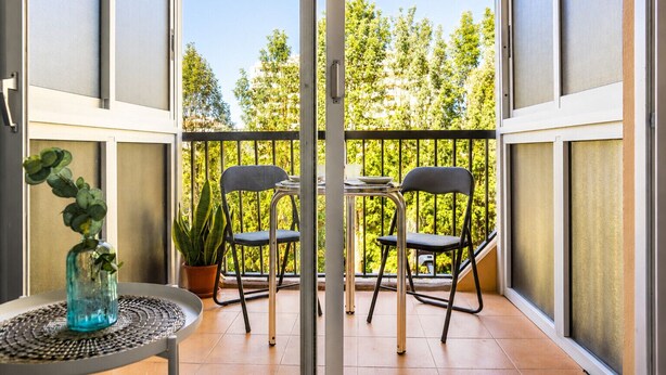 A balcony with two chairs and a table, a potted plant, and a view of trees.