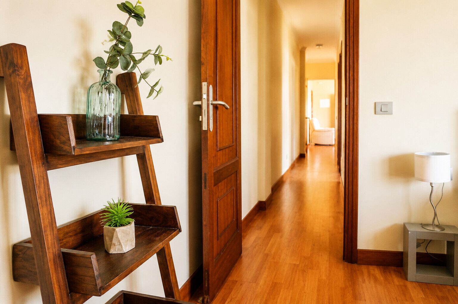Un interior de madera con un pasillo, una puerta, una mesita con una lámpara y una estantería con plantas.