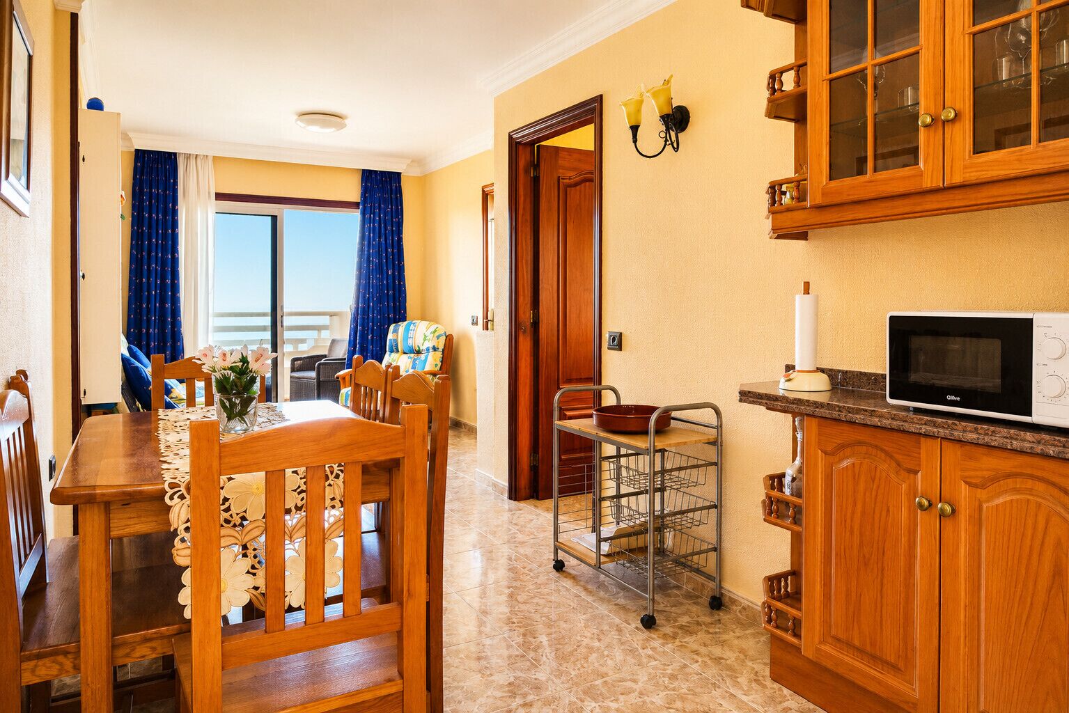 Un coin repas avec une table et des chaises, une cuisine avec des meubles en bois et un four à micro-ondes, et une vue sur un balcon aux rideaux bleus, accessible par une porte.
