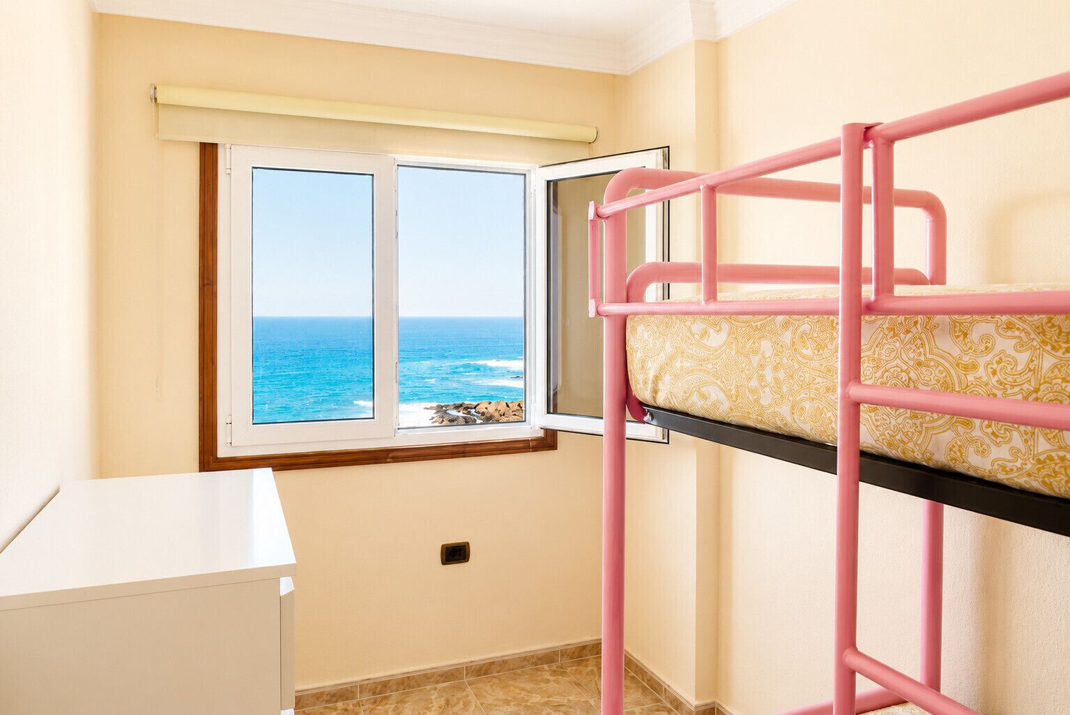 A bunk bed with a patterned mattress, a window with a view of the ocean, and a white desk.