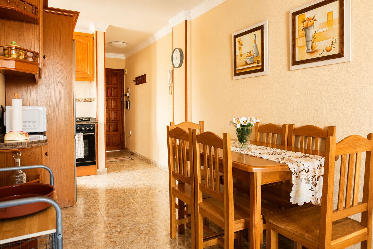 Une cuisine avec des meubles en bois, un four à micro-ondes et une table à manger avec des chaises. Il y a deux tableaux accrochés au mur.