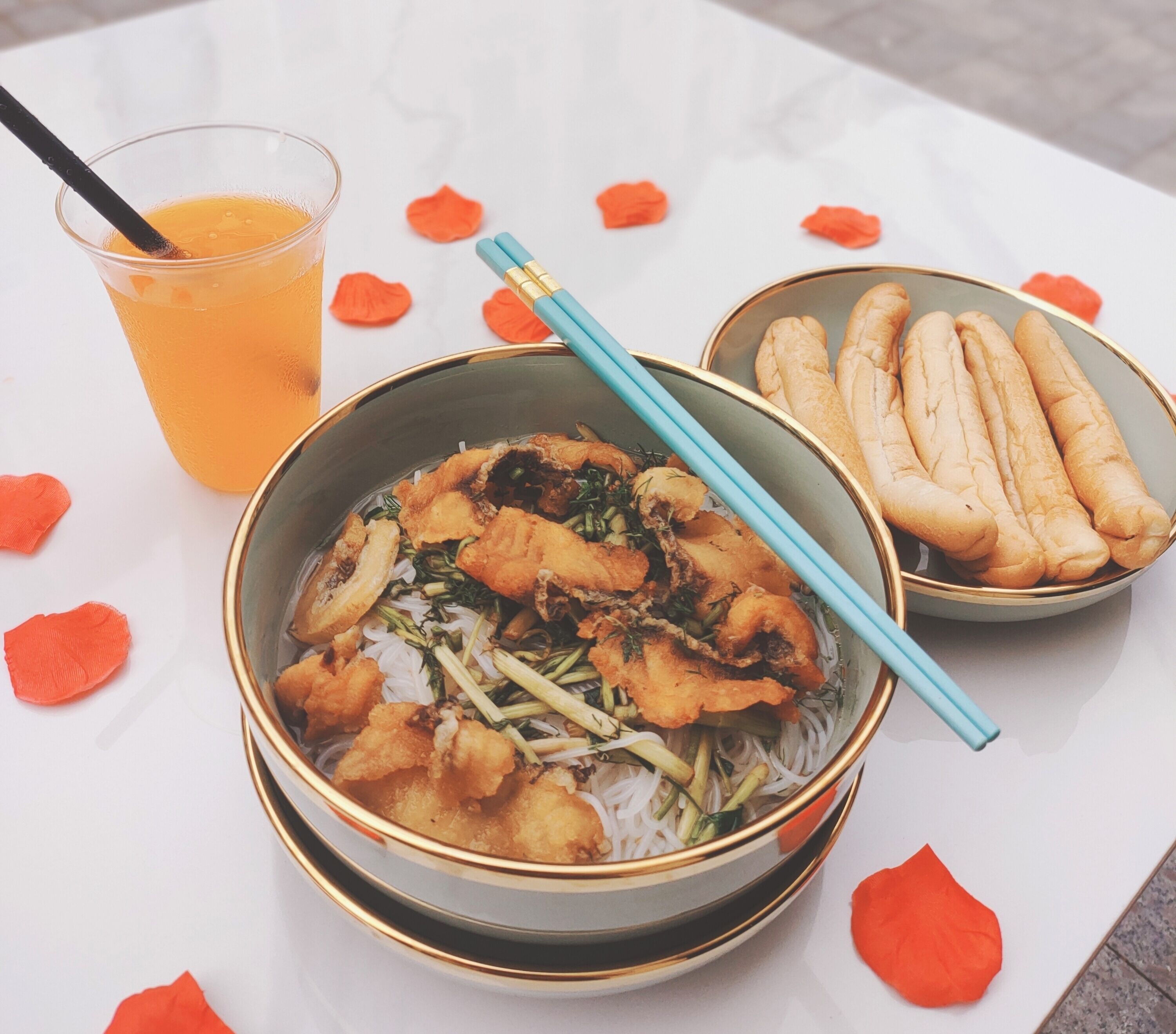 A bowl of noodles with fried items and vegetables, a glass of orange drink, and a bowl of breadsticks on a table with rose petals.