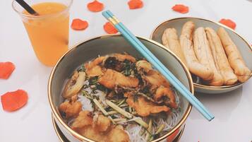 A bowl of noodles with fried items and vegetables, a glass of orange drink, and a bowl of breadsticks on a table with rose petals.