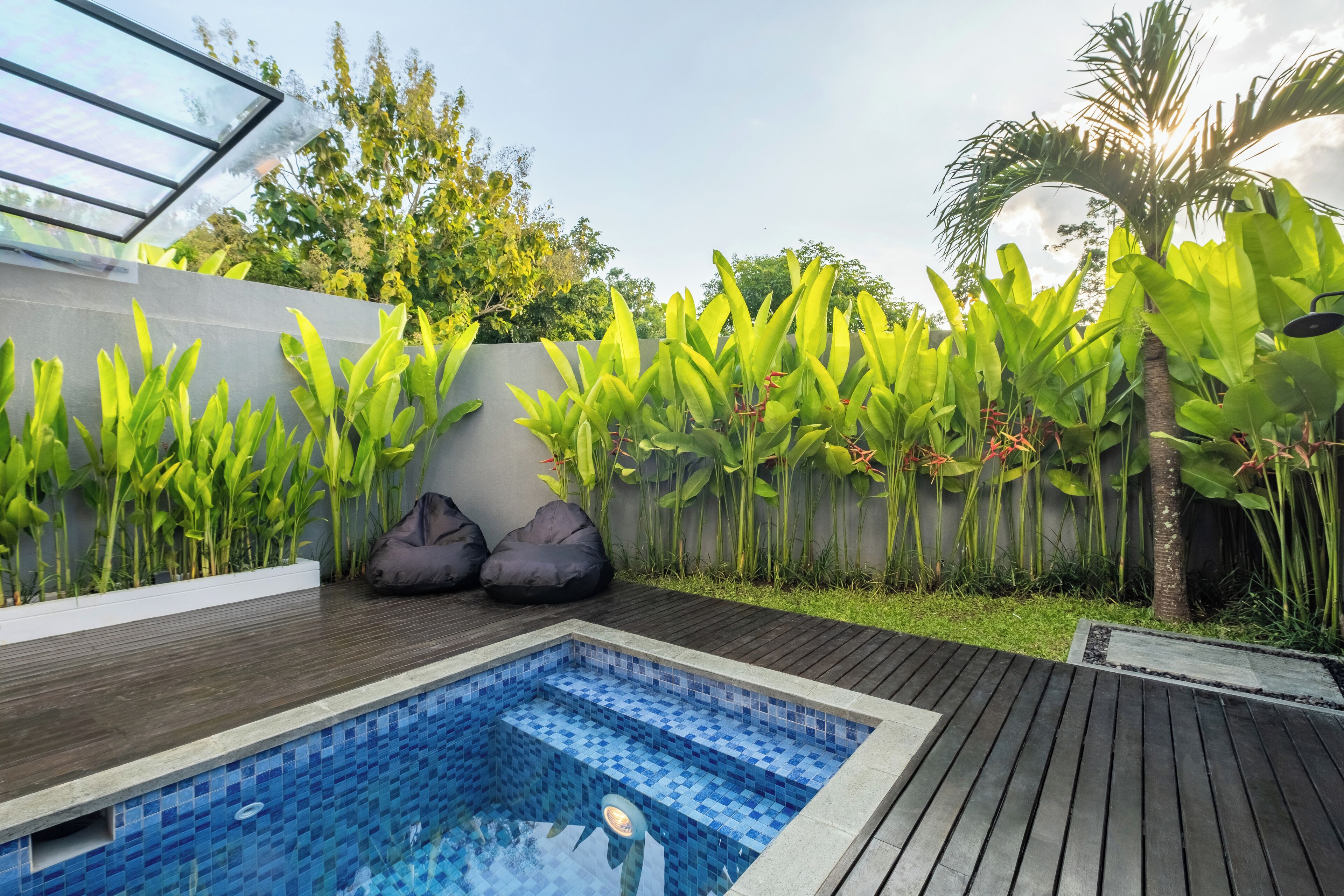 Ein Pool mit gefliestem Rand, umgeben von einer Holzterrasse und üppiger Vegetation, darunter Bananenpflanzen und eine Palme.