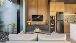 A modern living room with a sofa, a coffee table, and a TV. There is a kitchen area with a refrigerator and a microwave.