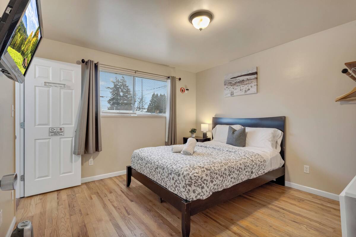 Un dormitorio con cama, ventana con cortinas, televisor montado en la pared, lámpara de noche y puerta con cartel.
