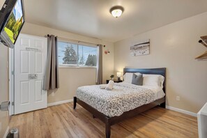 A bedroom with a bed, a window with curtains, a wall-mounted TV, a bedside lamp, and a door with a sign. - Brighton Guesthouse Private Rooms  (Seattle)