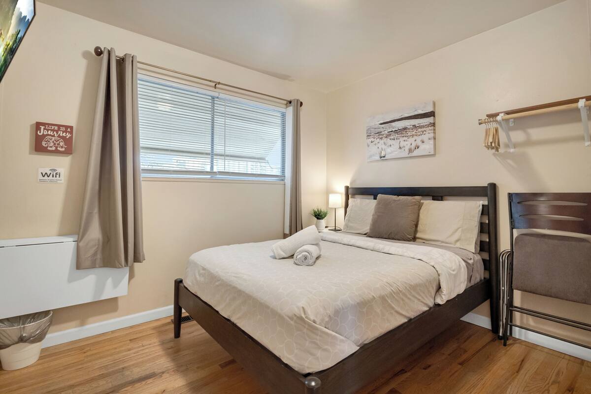 Una cama bien hecha con una colcha color beige y almohadas, una ventana con persianas, un cartel de WiFi montado en la pared y una pequeña planta en maceta sobre la mesita de noche.