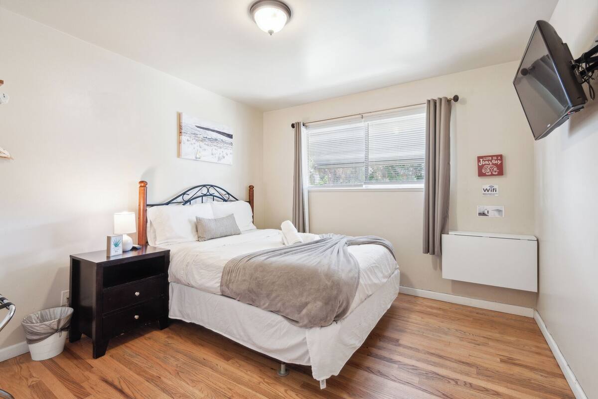 Un dormitorio con cama, mesita de noche, lámpara y televisor montado en la pared.