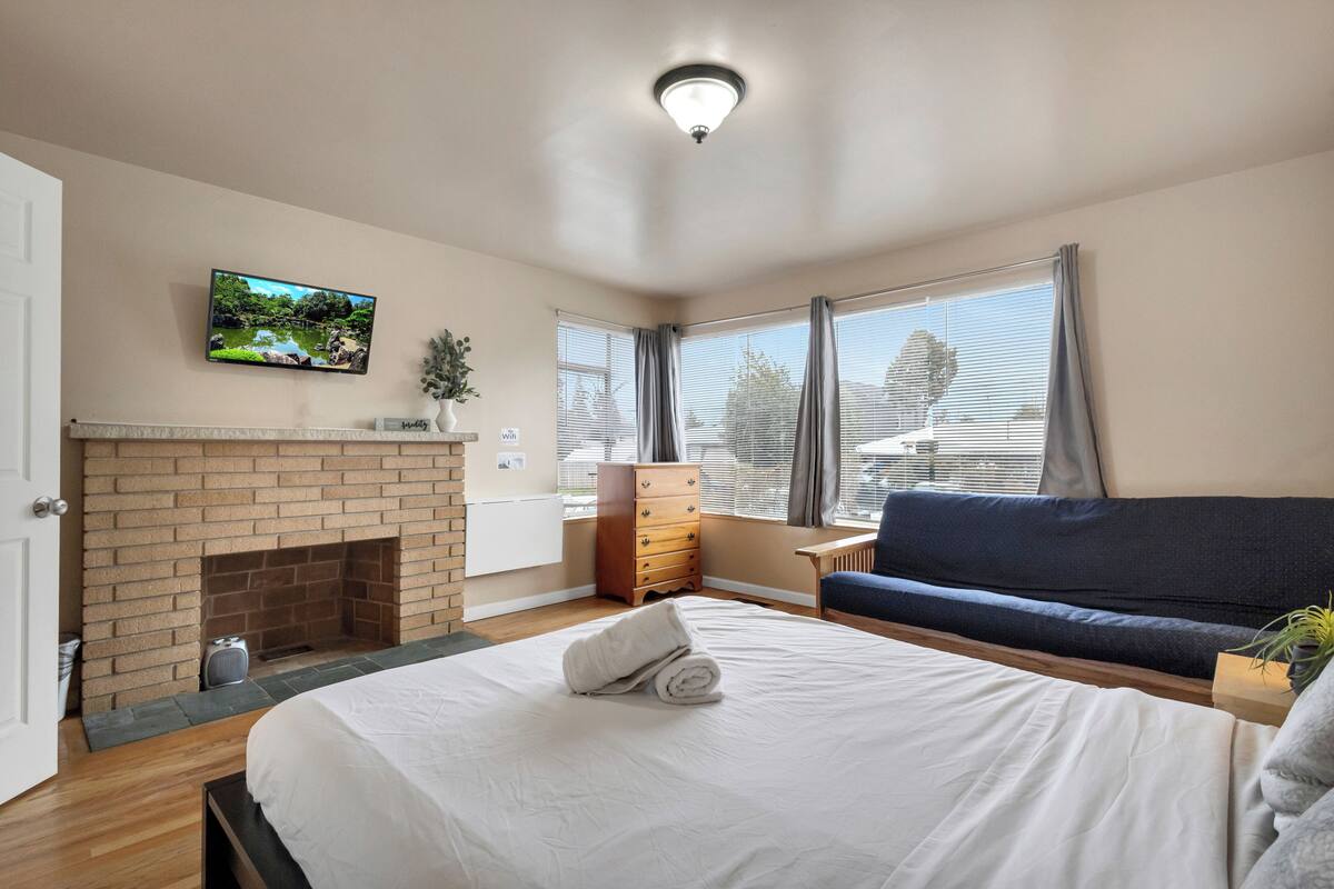 Una habitación de hotel con chimenea de ladrillo, una cama con ropa de cama blanca, un sofá azul, un televisor en la pared y una ventana con cortinas.