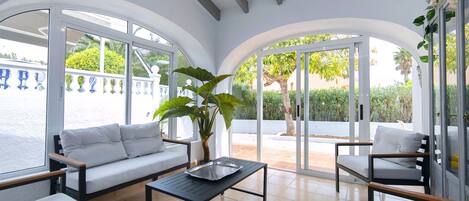 Une terrasse ensoleillée avec des canapés blancs, une table noire et une grande plante.