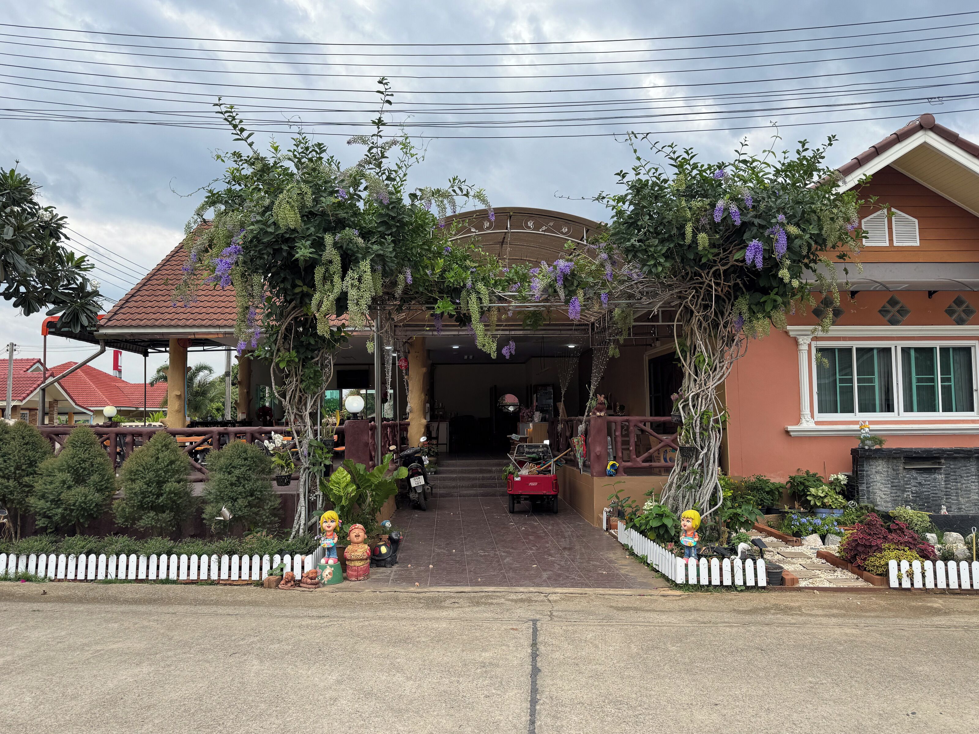 A covered entrance with a pergola adorned with hanging plants, a white picket fence, and a pathway leading to a building with a tiled roof.