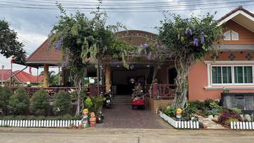 A covered entrance with a pergola adorned with hanging plants, a white picket fence, and a pathway leading to a building with a tiled roof.