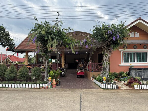 Una entrada cubierta con un pérgola decorada con plantas colgantes, una valla de piquetes blancos y un camino que conduce a un edificio con techo de tejas.