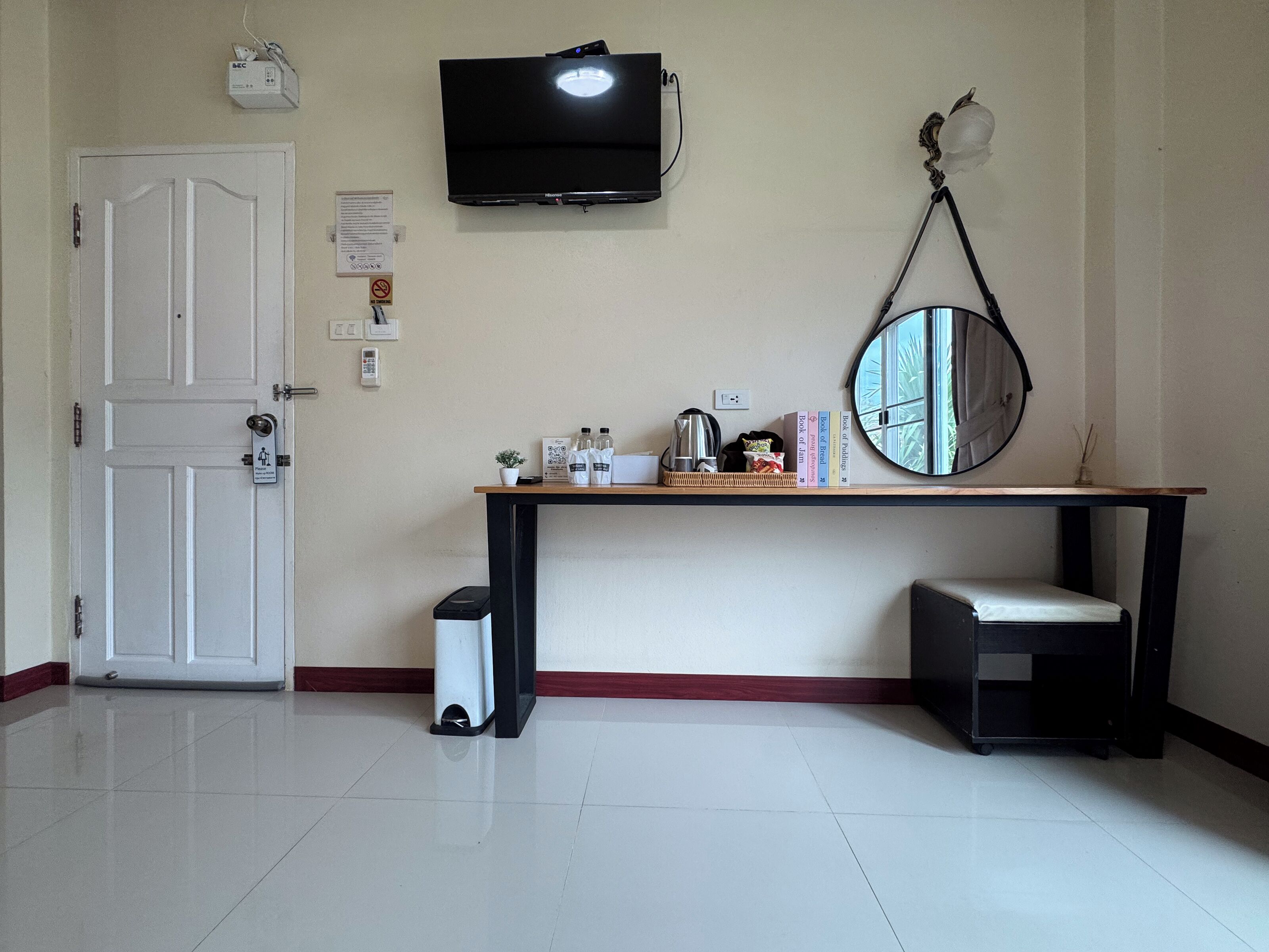 A clean, well-lit room with a white door, a small trash bin, a desk with a mirror, a television, and a shelf with books and a potted plant.