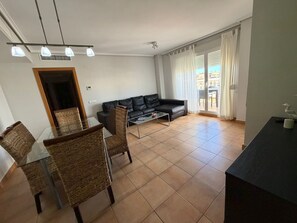 Ein Wohnzimmer mit einer schwarzen Ledersofa, einem Glastisch, Korbstühlen, einem Glastisch zum Essen und Blick durch eine Schiebetür ins Freie.