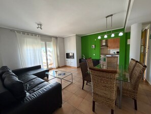 Una sala de estar con sofá, una mesa de centro de vidrio, sillas de mimbre, un televisor y una cocina verde al fondo.