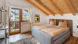 A bedroom with a large bed, wooden ceiling beams, a window with curtains, and a view of buildings outside. - Apartment 'Place In The Bavarian Mountains' with Mountain View, Private Garden and Wi-Fi (Schneizlreuth)