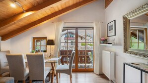 A dining area with a table and chairs, a window with curtains, a radiator, and a mirror on the wall. - Apartment 'Place In The Bavarian Mountains' with Mountain View, Private Garden and Wi-Fi (Schneizlreuth)