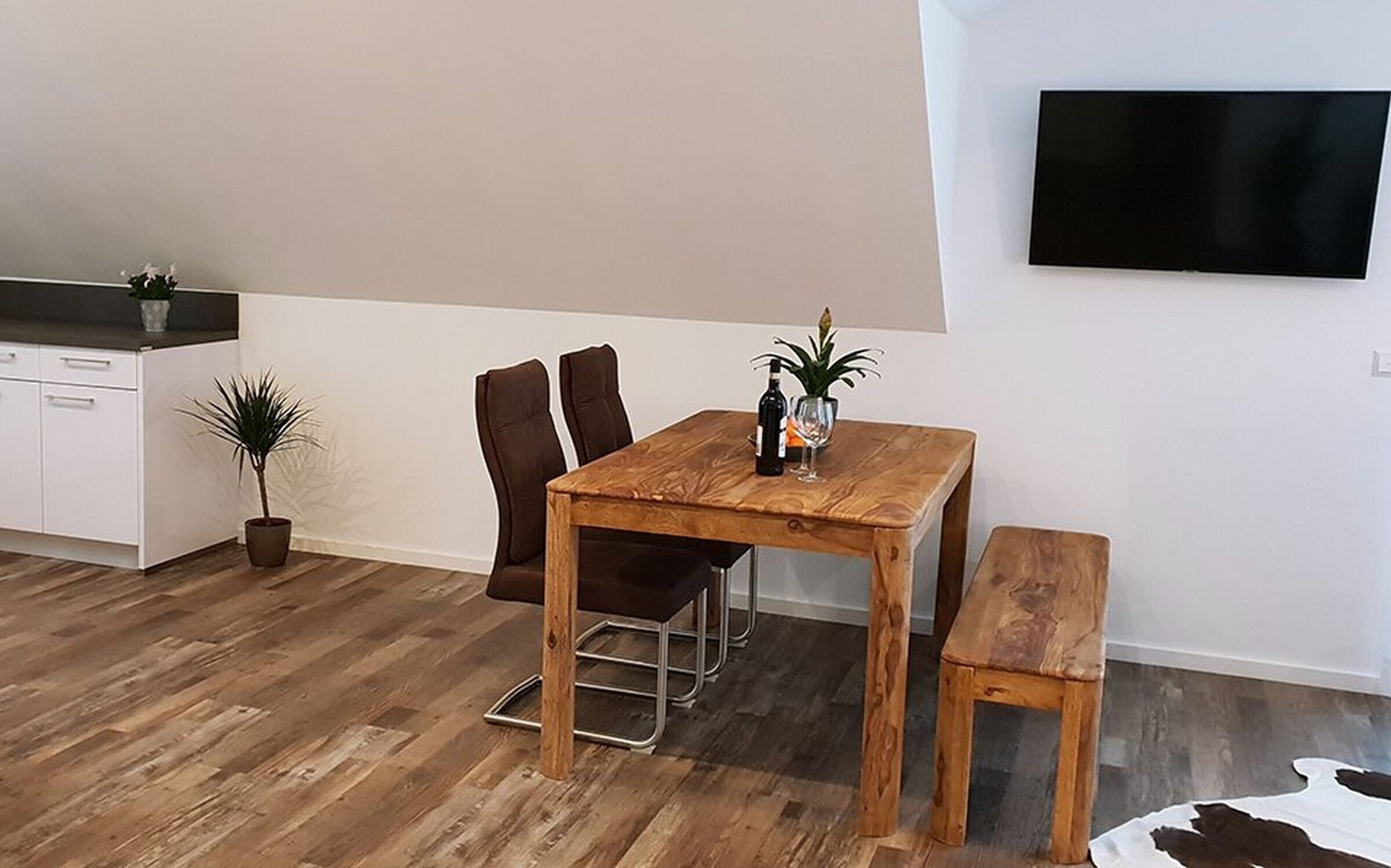 Un comedor con una mesa de madera, dos sillas, un banco, una planta en maceta, una botella de vino y una copa.
