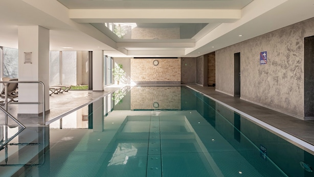 A modern indoor swimming pool with clear water, a glass ceiling, and a stone wall.