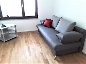 A grey sofa with red and grey cushions, a small white side table, and a window with blinds. - Apartment 'Enzblick' with Balcony and Wi-Fi (Bad Wildbad)