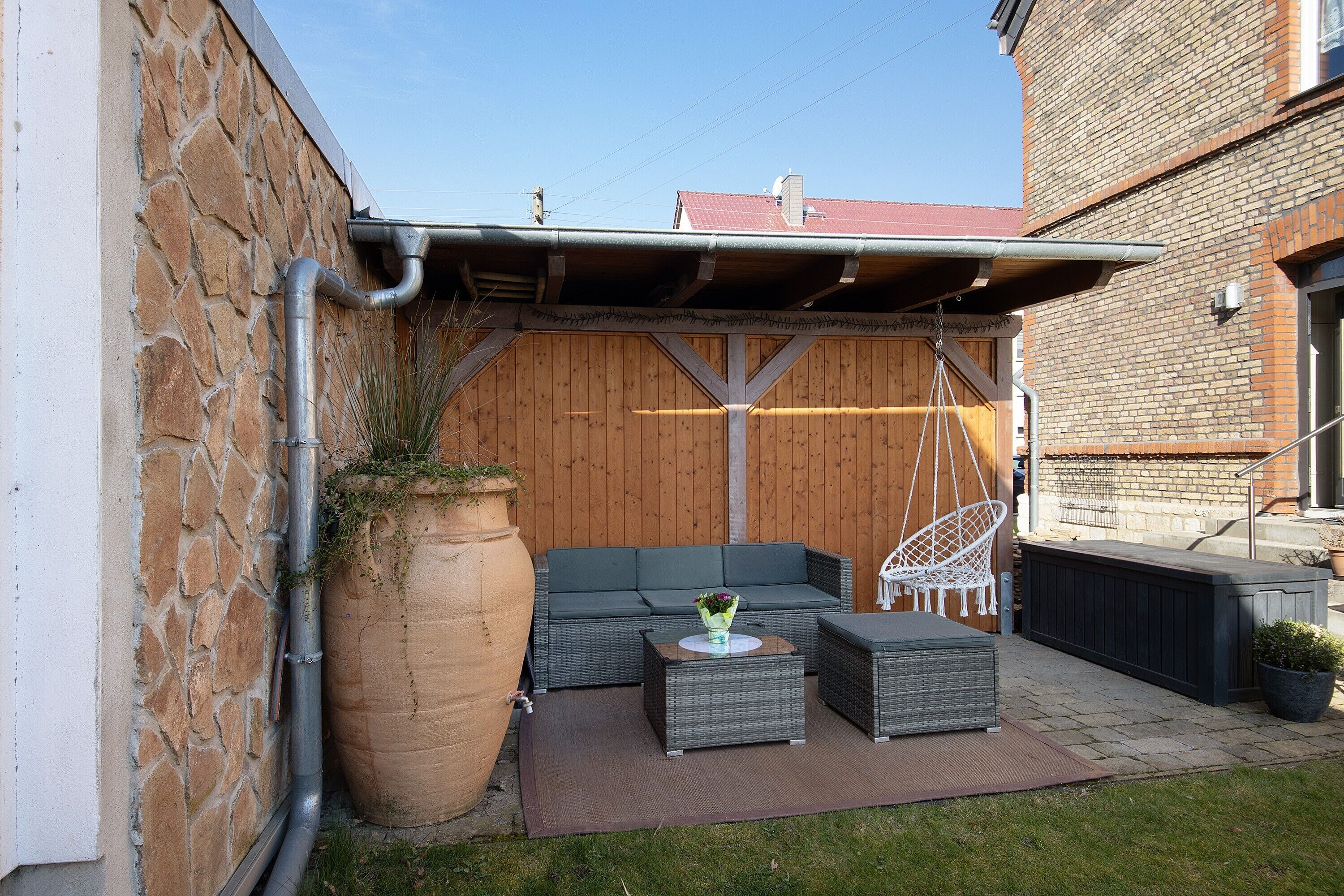 A patio with a stone wall, a wooden fence, a grey sofa, a white chair, and a large planter.