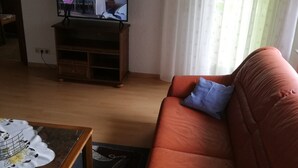 Una sala de estar con una televisión sobre un mueble de madera, un sofá rojo con un cojín azul y una mesa con estampado.