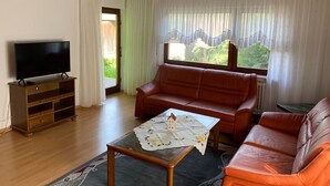 Una sala de estar con televisor, sofá, una mesa con un modelo de casa y una ventana con cortinas.