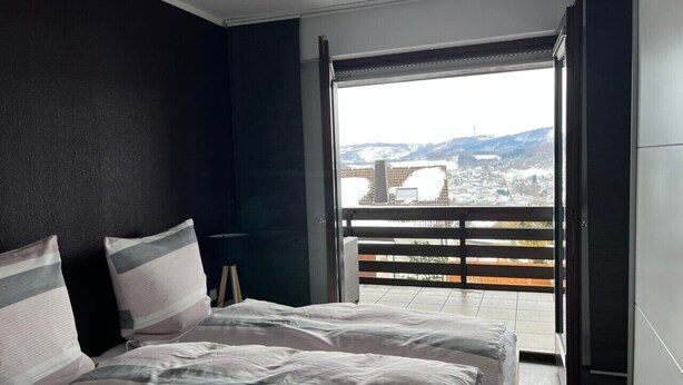 A bedroom with a large bed, a nightstand, and a view of a cityscape through an open door. - Apartment "Lavender", 110 sqm with Balcony (Herscheid)