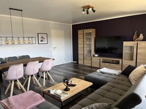 Uma sala de estar moderna com uma mesa de jantar de madeira, cadeiras estofadas, um sofá, uma mesa de centro, uma TV e armários embutidos.