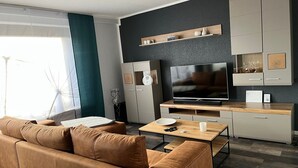 A modern living room with a sofa, coffee table, TV, and built-in cabinets. - Haus Blumenglück "Lavender and Dandelion" – Two Apartments for up to 11 Guests (Herscheid)