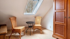 Una habitación con techo inclinado, una ventana, dos sillas de mimbre, una pequeña mesa redonda y un aparador de madera.