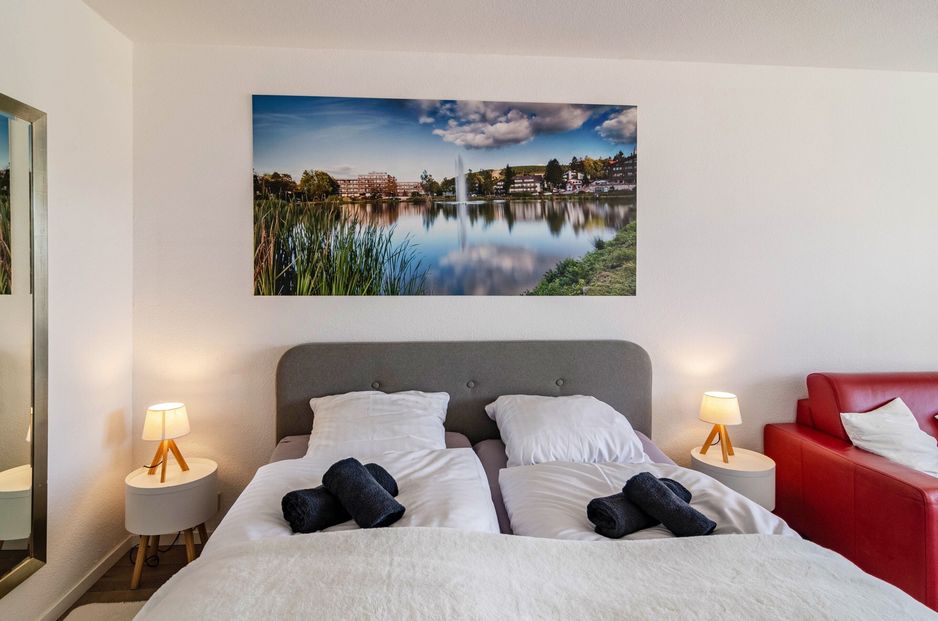 A hotel room with a bed, two bedside tables with lamps, a red chair, and a large landscape painting on the wall.