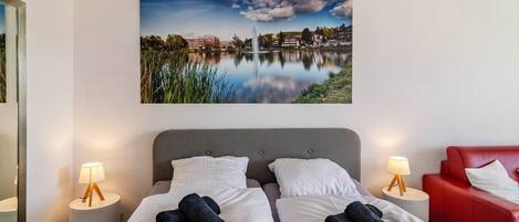 A hotel room with a bed, two bedside tables with lamps, a red chair, and a large landscape painting on the wall.