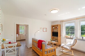 Un salon confortable avec un canapé, un fauteuil, une petite table et un téléviseur. Il y a une fenêtre avec des rideaux et une vue sur la verdure à l’extérieur.