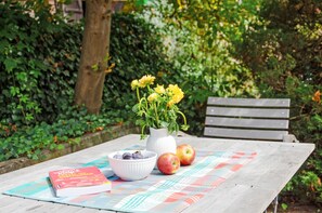 A wooden table with a placemat, a white vase with yellow flowers, a bowl of plums, and two apples.