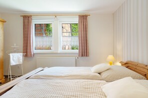 Quarto de hotel com duas camas, janela com cortinas, radiador, mesa com abajur e um porta-toalhas.
