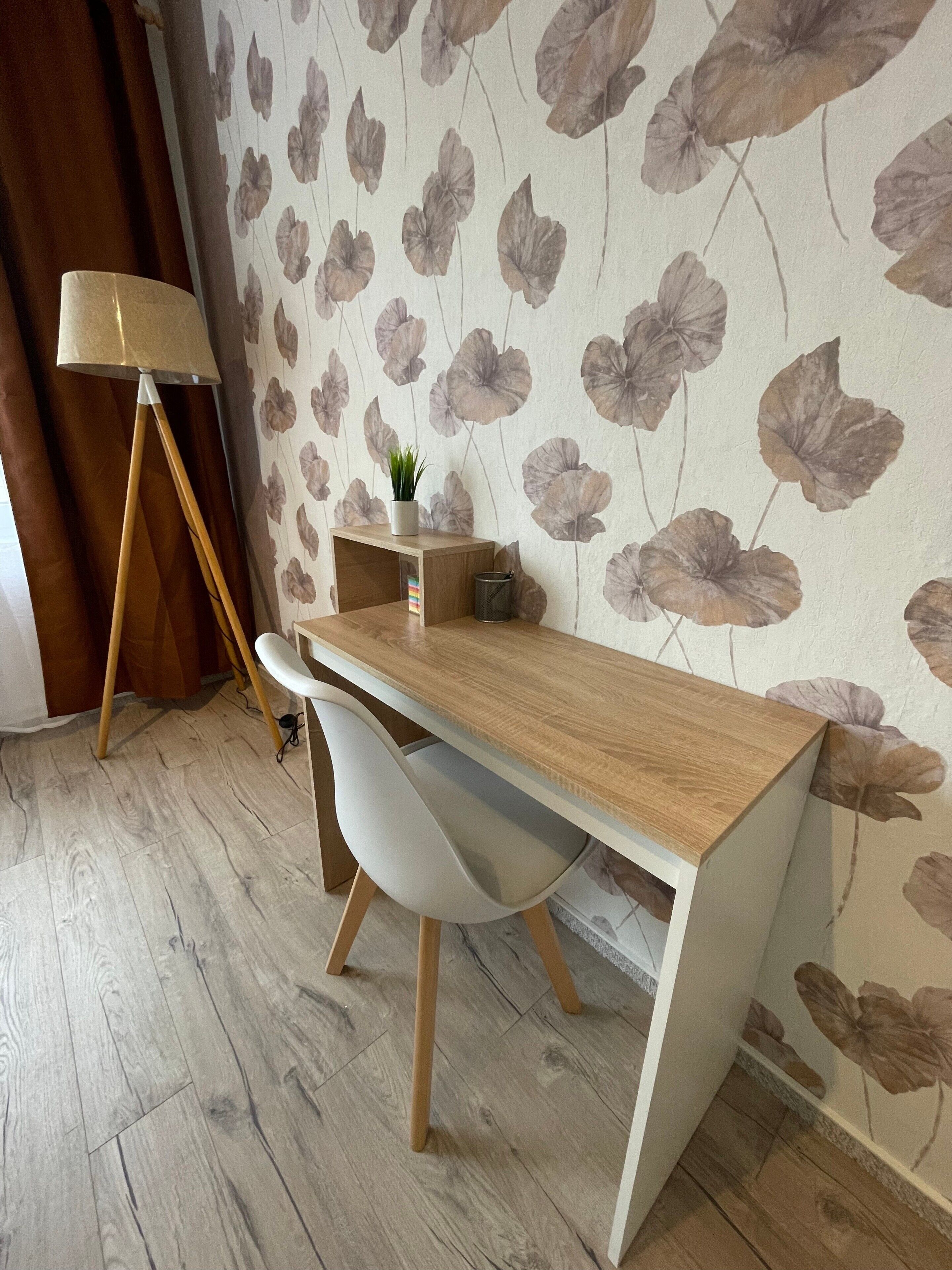 A wooden desk with a white chair, a small plant, and a lamp in a room with floral wallpaper and wooden flooring.