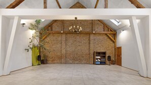 A spacious room with exposed brick walls, a high ceiling with wooden beams, a chandelier, and a fireplace. - Finca 'Gottsdorf' with Private Terrace and Shared Garden (Nuthe-Urstromtal)
