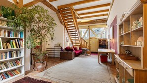 A spacious loft with exposed wooden beams, a central staircase, a large bookshelf, a potted plant, and a cozy seating area. - Finca 'Gottsdorf' with Private Terrace and Shared Garden (Nuthe-Urstromtal)