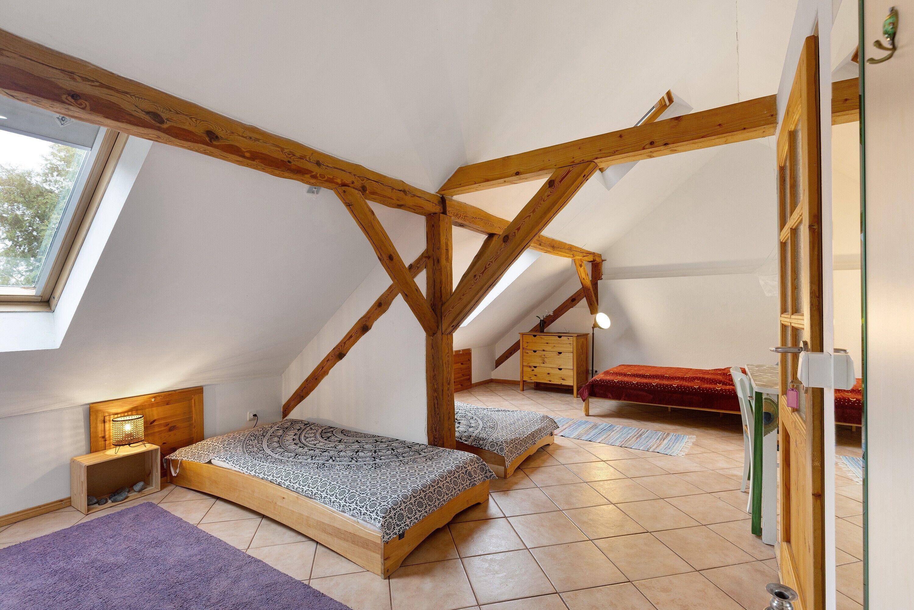 Ein Schlafzimmer im Spitzboden mit schrägen Wänden, Holzbalken, zwei Betten, einer Kommode und einem Fenster.